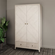 The Diletta 2 Door 2 Drawer Wardrobe in light wood offers elegant bedroom storage, standing against a beige wall on a wooden floor beside a green potted plant in a metal stand.