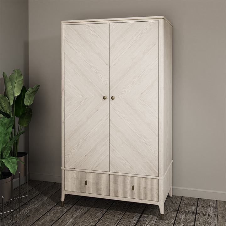 The Diletta 2 Door 2 Drawer Wardrobe in light wood offers elegant bedroom storage, standing against a beige wall on a wooden floor beside a green potted plant in a metal stand.