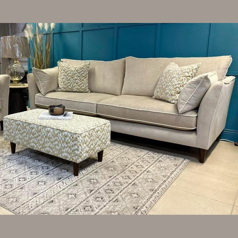 The Irish-made Layla Grand Sofa with washable aquaclean fabrics displays patterned cushions by a teal wall; a matching ottoman rests on a light gray rug, accented with a lamp, decorative grass, and magazines.
