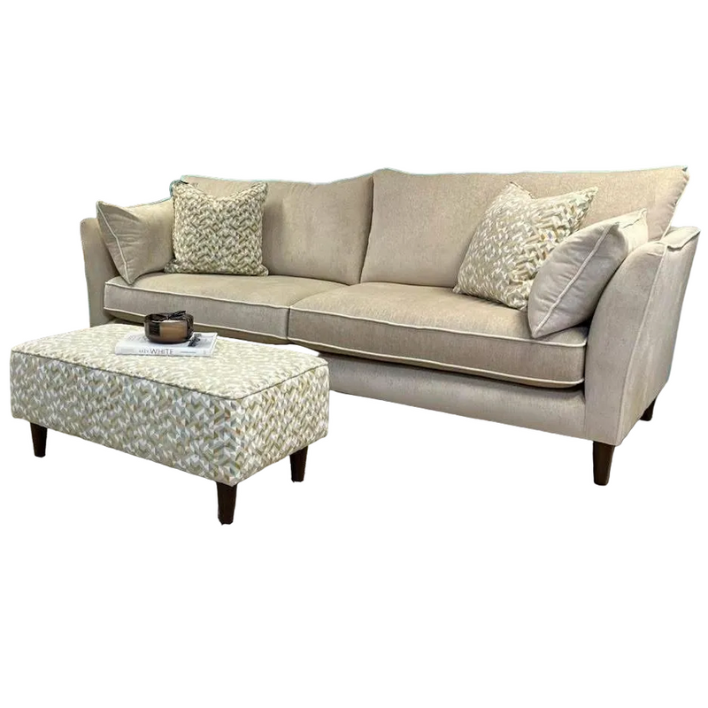 The Layla Grand Sofa in beige aquaclean upholstery, made in Ireland, includes two patterned throw pillows. A matching ottoman on dark wood legs holds a tray with a cup and magazine.