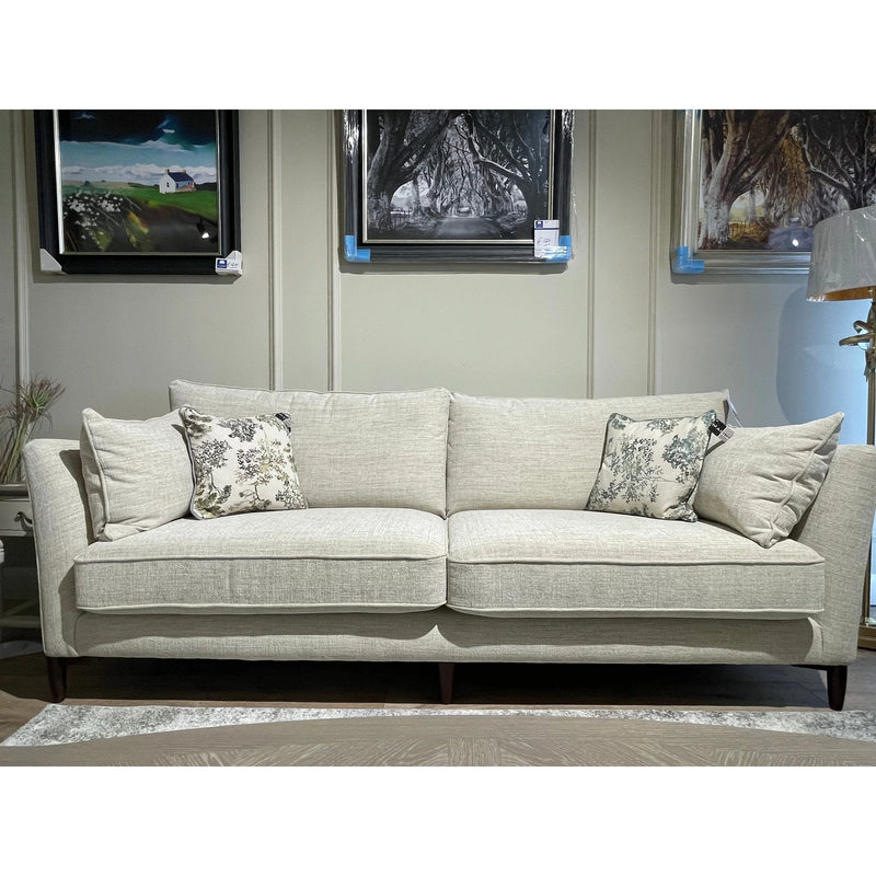 The Layla Grand Sofa in light beige, Irish-made with washable Aquaclean fabric, includes two printed throw pillows and sits against a gray paneled wall beneath framed art in a stylish living room.