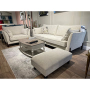 A modern living room display showcases the Irish-made Layla Grand Sofa in light beige washable aquaclean fabric with a matching ottoman, a rectangular coffee table, light gray rug on wooden floors, and shelves with decor in the background.