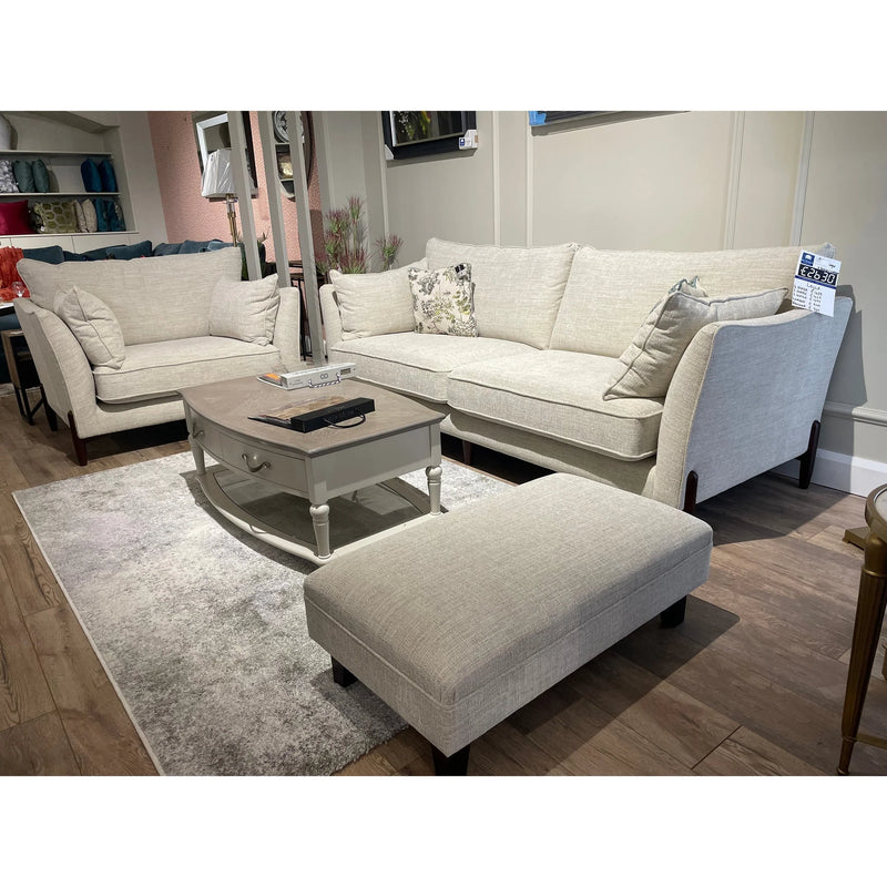 A modern living room display showcases the Irish-made Layla Grand Sofa in light beige washable aquaclean fabric with a matching ottoman, a rectangular coffee table, light gray rug on wooden floors, and shelves with decor in the background.
