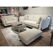 A modern living room display showcases the Irish-made Layla Grand Sofa in light beige washable aquaclean fabric with a matching ottoman, a rectangular coffee table, light gray rug on wooden floors, and shelves with decor in the background.