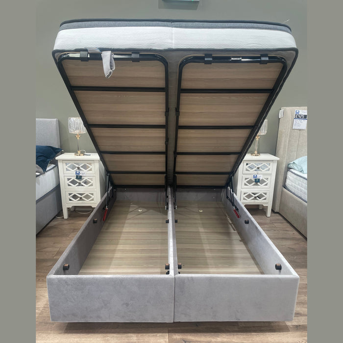 The Bella Ottoman Bed, upholstered in gray, features a lift-up mattress platform revealing two spacious storage compartments underneath. It is placed between two white nightstands with lamps.