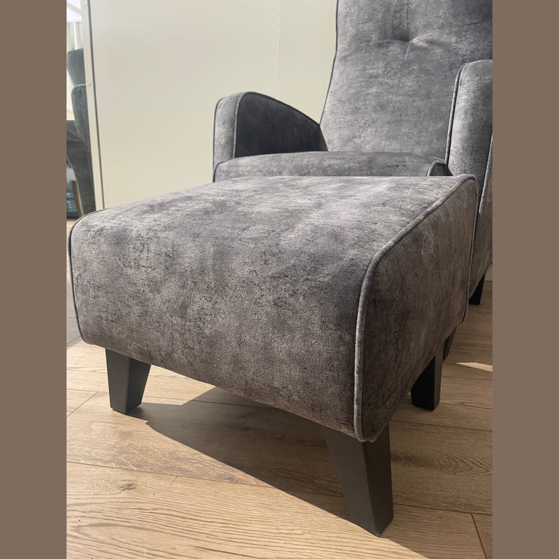 The Kelvin Chair, featuring gray upholstery, black angled legs, and a matching footstool, sits on a light wood floor with sunlight streaming in from the left. Its sleek, minimalist design complements any modern space.