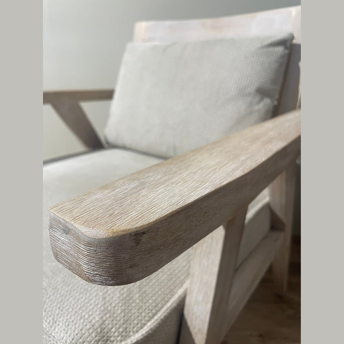 Close-up shot highlighting the texture of a distressed or wire-brushed light wood armchair frame, featuring a rounded armrest and a neutral beige fabric cushion in the background.