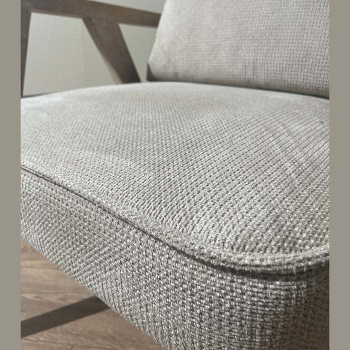 A close-up of the Willow Chair with light gray beige upholstery, wooden armrests, and textured fabric on a cushioned seat, set on a wooden floor against a plain light wall.