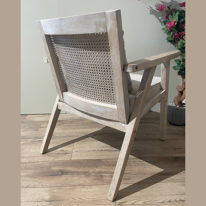 The Willow Chair, featuring a cane mesh backrest and beige upholstered seat, sits on a wooden floor with a pot of pink flowers and green leaves in the background.