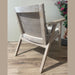The Willow Chair, featuring a cane mesh backrest and beige upholstered seat, sits on a wooden floor with a pot of pink flowers and green leaves in the background.