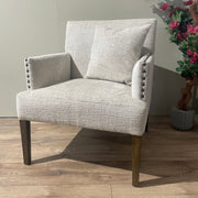 The Sylvia Chair has light beige upholstery, dark wooden legs, beaded arm detailing, and a matching cushion. It’s placed on a wooden floor next to a green plant and a faux flowering tree.