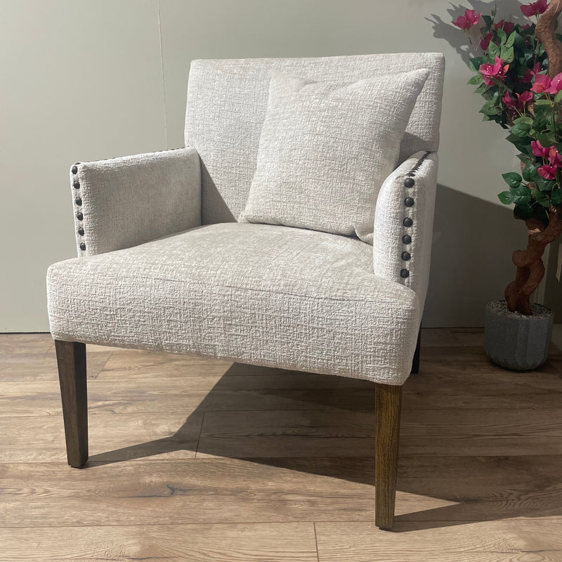 The Sylvia Chair has light beige upholstery, dark wooden legs, beaded arm detailing, and a matching cushion. It’s placed on a wooden floor next to a green plant and a faux flowering tree.