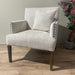 The Sylvia Chair has light beige upholstery, dark wooden legs, beaded arm detailing, and a matching cushion. It’s placed on a wooden floor next to a green plant and a faux flowering tree.
