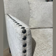 Close-up of the Sylvia Chair with light beige upholstery and black beaded details on the armrest. Includes matching cushion; blurred plant in background.