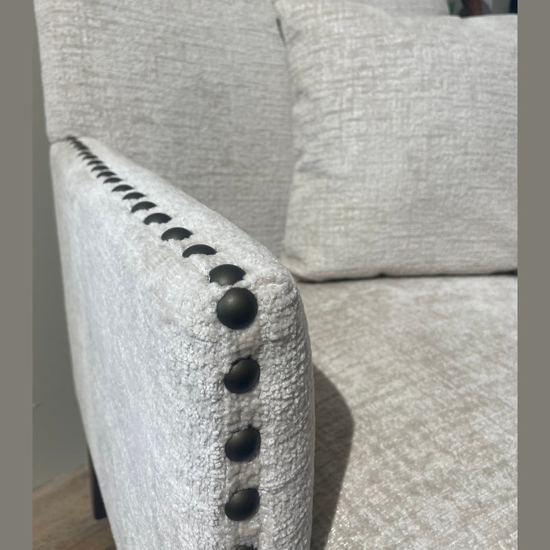 Close-up of the Sylvia Chair with light beige upholstery and black beaded details on the armrest. Includes matching cushion; blurred plant in background.