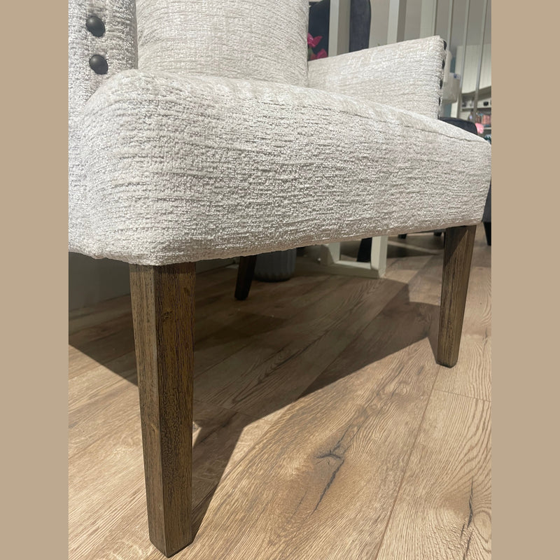 A close-up view of the Sylvia Chair highlights its light beige textured upholstery, dark wooden legs, and elegant beaded trim, shown indoors on a light wood floor.