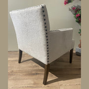 The Sylvia Chair showcases light beige upholstery, dark wooden legs, and black beaded trim, placed on a wood floor near a sunlit window with pink flowers.