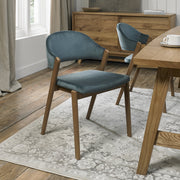 A Camden Dining Chair with a blue velvet seat and backrest is placed on a light-patterned rug in a dining room featuring wooden floors, a matching table, and a sideboard in the background.