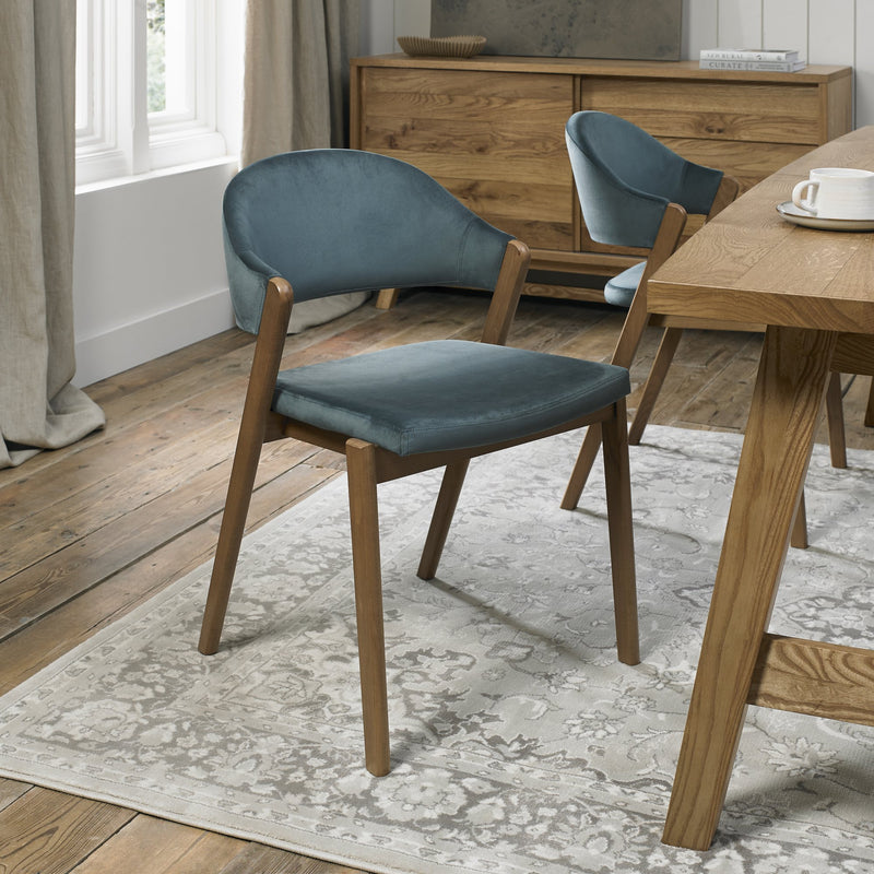 A Camden Dining Chair with a blue velvet seat and backrest is placed on a light-patterned rug in a dining room featuring wooden floors, a matching table, and a sideboard in the background.