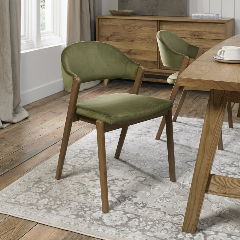 A Camden Dining Chair in olive green velvet adds style to a modern dining room with wooden floors, a wood table, sideboard, and a patterned light gray rug.