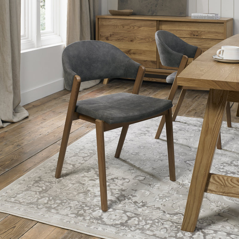 A modern dining room with a wooden floor, light patterned rug, and Camden Dining Chairs with curved backs and dark gray cushions surrounds a wooden table. Beige curtains frame the window and a wooden sideboard is in the background.
