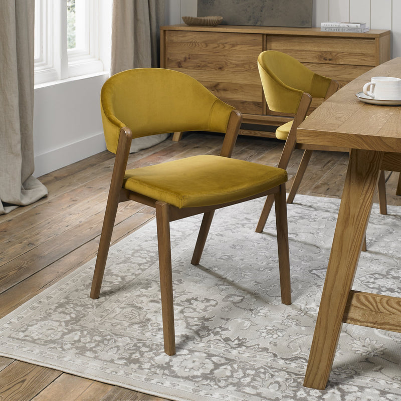 The Camden Dining Chair in mustard yellow velvet with wooden legs is placed on a patterned rug, surrounded by vibrant colors, natural light, and wooden furniture in the dining room.