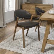 A Camden Dining Chair with a curved black leather backrest and seat is placed on a patterned rug beside a wooden dining table in a bright room with wood floors and colorful decor, with a sideboard in the background.