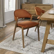 A Camden Dining Chair in burnt orange velvet sits on a patterned gray rug in a bright room with wood floors, a light wood dining table, and a matching sideboard.
