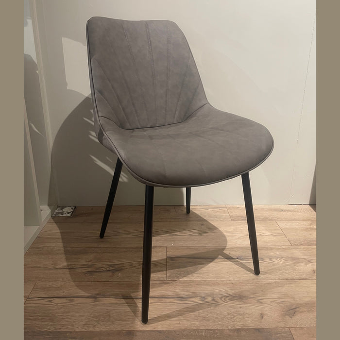 The Lilly Dining Chair features modern gray faux leather and angled black legs, shown on a wooden floor against a plain light-colored wall.