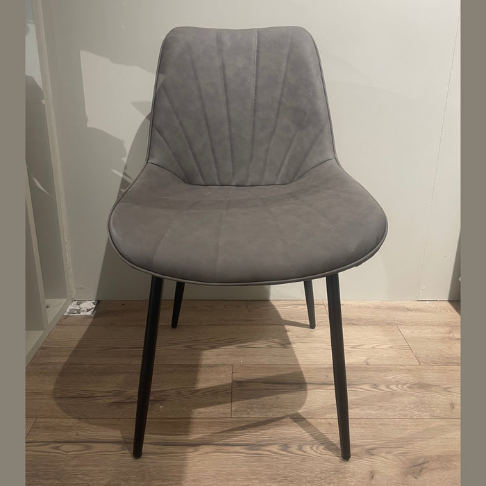 The Lilly Dining Chair, with black metal legs, sits on a light wood floor against a pale wall. This modern gray chair features a smooth, subtly curved back and seat with delicate stitched lines for a contemporary look.