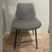 The Lilly Dining Chair, with black metal legs, sits on a light wood floor against a pale wall. This modern gray chair features a smooth, subtly curved back and seat with delicate stitched lines for a contemporary look.