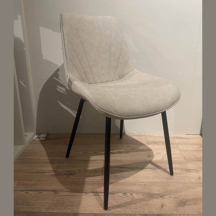 The Lilly Dining Chair, featuring light beige upholstery and angled black legs, is placed on a wooden floor against a plain white wall.