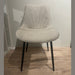 The Lilly Dining Chair features beige upholstery, angled black legs, and a curved backrest with subtle stitching—perfect as a modern accent piece for any space. It sits stylishly on light wood flooring against an off-white wall.