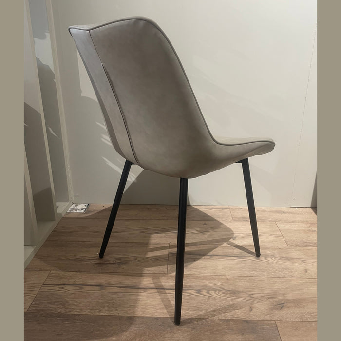 The Lilly Dining Chair features a modern design with light gray faux leather, a curved backrest, and black metal legs, standing on a wooden floor against a plain light-colored wall.
