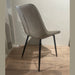 The Lilly Dining Chair features a modern design with light gray faux leather, a curved backrest, and black metal legs, standing on a wooden floor against a plain light-colored wall.