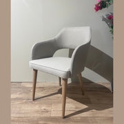 The Turim Dining Chair, featuring light gray upholstery and wooden legs, sits on a wooden floor beside a wall and a pink-flowered plant, blending contemporary comfort with retro-inspired style.