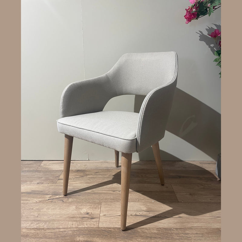 The Turim Dining Chair, featuring light gray upholstery and wooden legs, sits on a wooden floor beside a wall and a pink-flowered plant, blending contemporary comfort with retro-inspired style.
