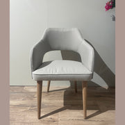 The Turim Dining Chair, featuring light gray upholstery and wooden legs, stands on a wooden floor against a plain light wall. A floral arrangement in the corner complements the chair’s sleek, retro-inspired design.
