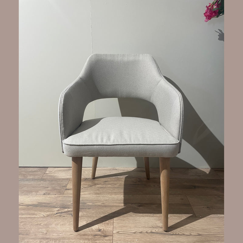 The Turim Dining Chair, featuring light gray upholstery and wooden legs, stands on a wooden floor against a plain light wall. A floral arrangement in the corner complements the chair’s sleek, retro-inspired design.