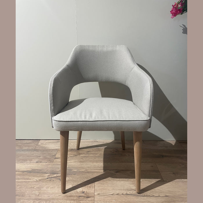 The Turim Dining Chair, featuring light gray upholstery and wooden legs, stands on a wooden floor against a plain light wall. A floral arrangement in the corner complements the chair’s sleek, retro-inspired design.