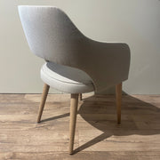The Turim Dining Chair features light-gray upholstery, wooden legs, and a curved back with an open lower back. It is shown at an angle on a wooden floor against a plain, light-colored wall to highlight its modern design.