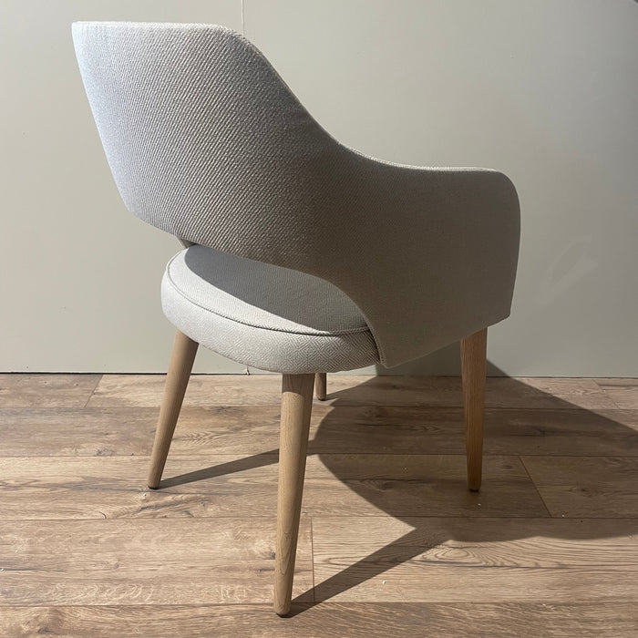 The Turim Dining Chair features light-gray upholstery, wooden legs, and a curved back with an open lower back. It is shown at an angle on a wooden floor against a plain, light-colored wall to highlight its modern design.