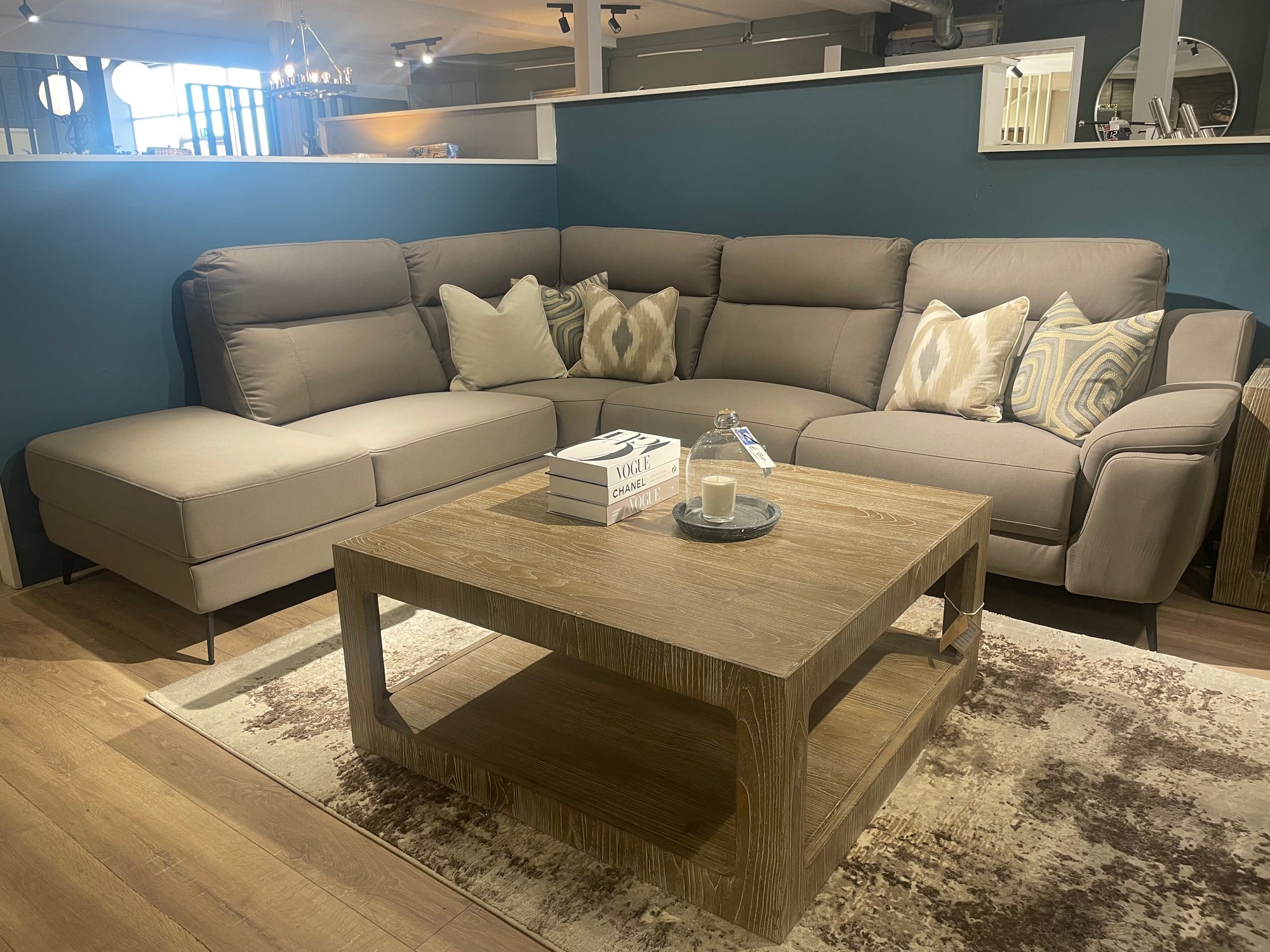 Furniture showroom view of a tan sectional couch and a square, open-frame, light-colored wood coffee table with styled accessories.