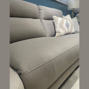 A close-up of the Chloe Corner Group, a modern gray sofa with cushioned backrests, shown with two decorative pillows—one in a taupe and beige pattern, the other with blue and gray stripes—with a lamp in the background.