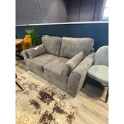 The Roma Sofabed, a gray upholstered compact sofabed with plush cushions and armrests, sits next to a light gray chair, a plant, and a table on a patterned beige rug in a modern room with teal walls.
