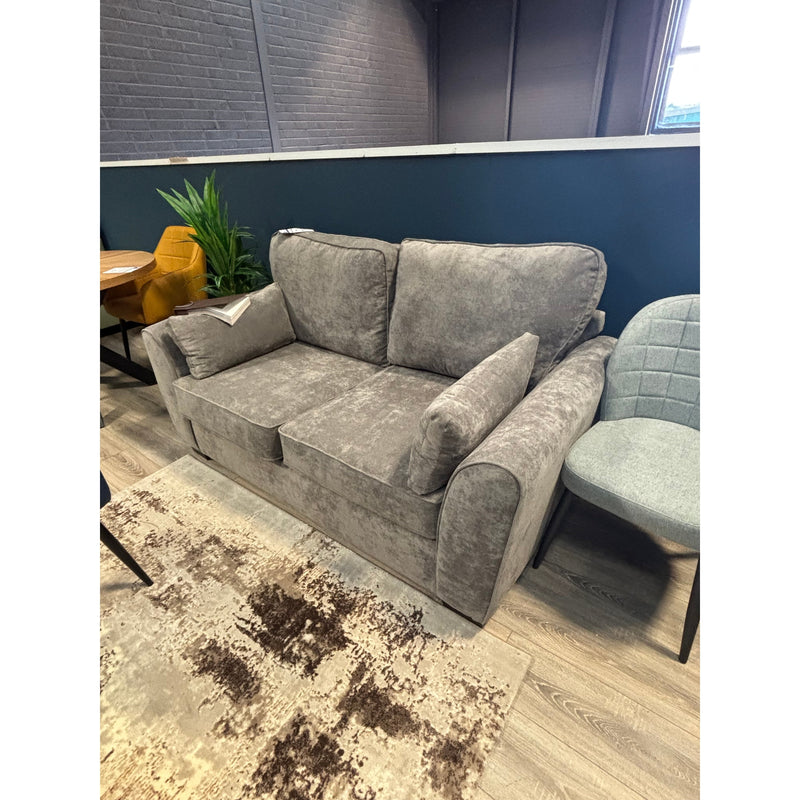 The Roma Sofabed, a gray upholstered compact sofabed with plush cushions and armrests, sits next to a light gray chair, a plant, and a table on a patterned beige rug in a modern room with teal walls.