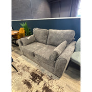 Small, comfortable gray loveseat with deep seating, photographed in a furniture store against a dark blue wall and brick background.