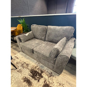 The Roma Sofabed, in gray upholstery with matching cushions, is placed in a modern room featuring wood floors, a patterned rug, a yellow chair, green plants, and contrasting dark blue and brick walls.