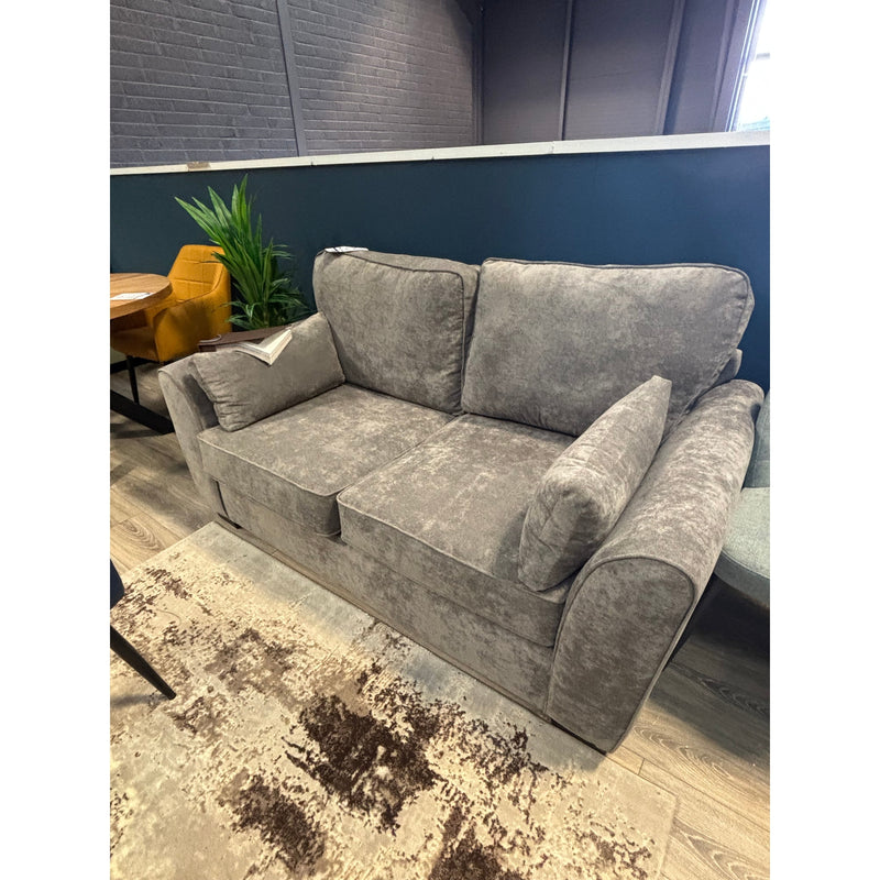 The Roma Sofabed, in gray upholstery with matching cushions, is placed in a modern room featuring wood floors, a patterned rug, a yellow chair, green plants, and contrasting dark blue and brick walls.