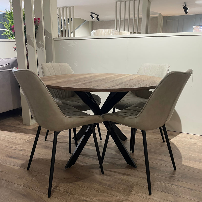The Elson Round Dining Table with black X legs is paired with four light beige cushioned chairs on a wooden floor in a bright, modern room.